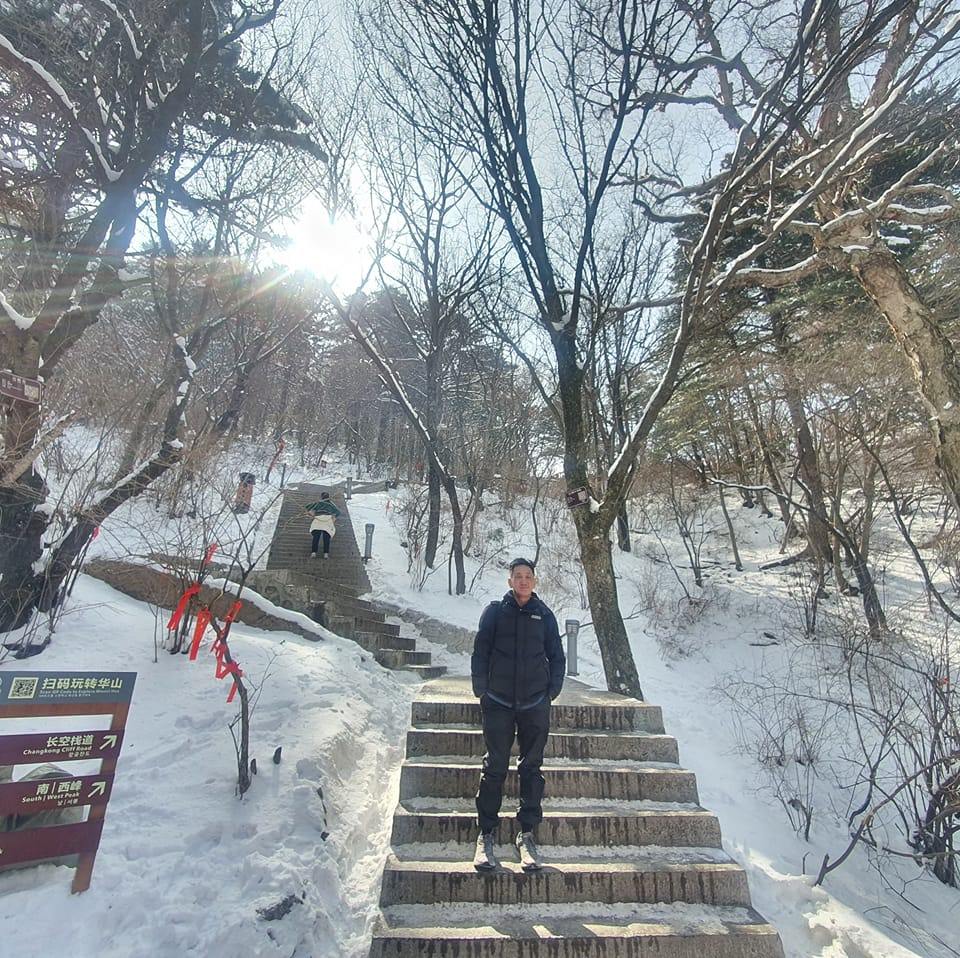 Anthony K. H. Tung standing on the snow-covered stone steps of Mount Hua (Huashan) in Shaanxi, China, during winter, with bare trees and mountain peaks behind.
